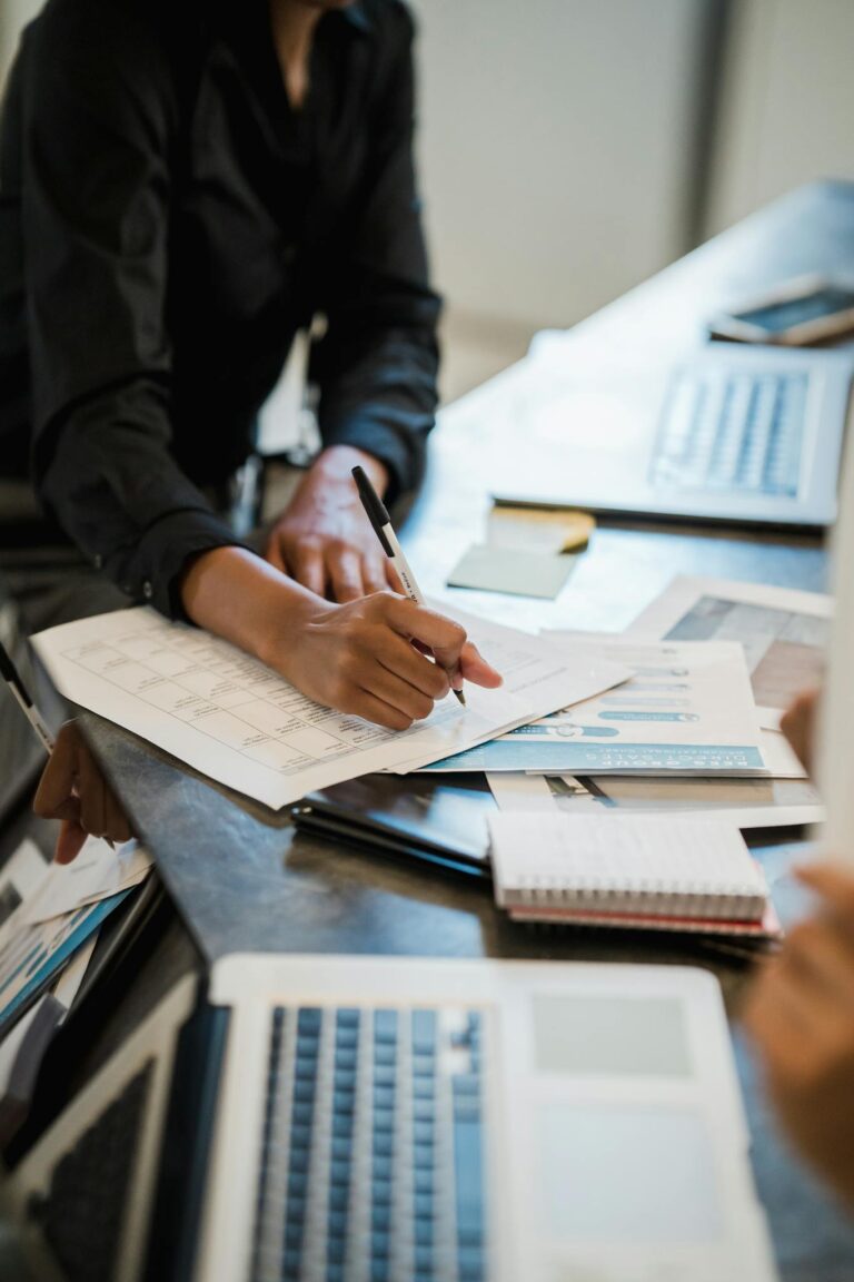 Close-up of a professional writing on documents in a busy office environment with laptops on a desk.
