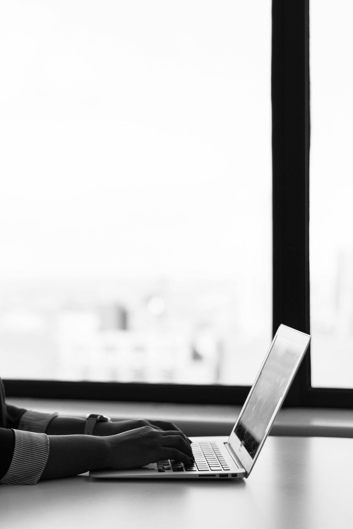Hands typing on a laptop by a window in a modern setting, emphasizing productivity and isolation.